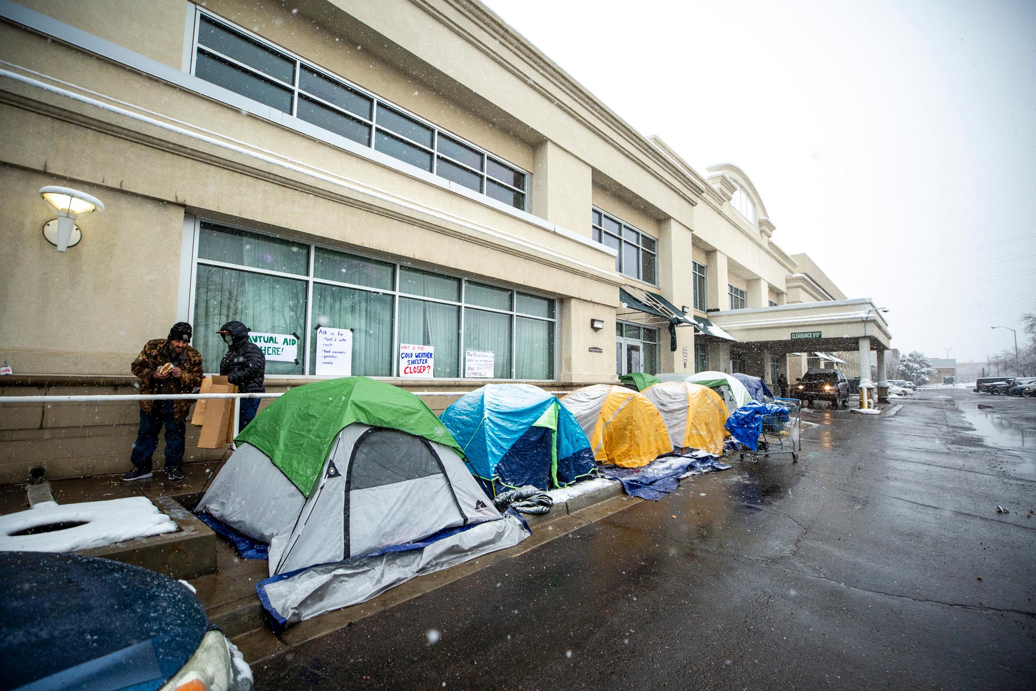 Colorful tents are set up in front of a beige building as snow falls in the foreground.