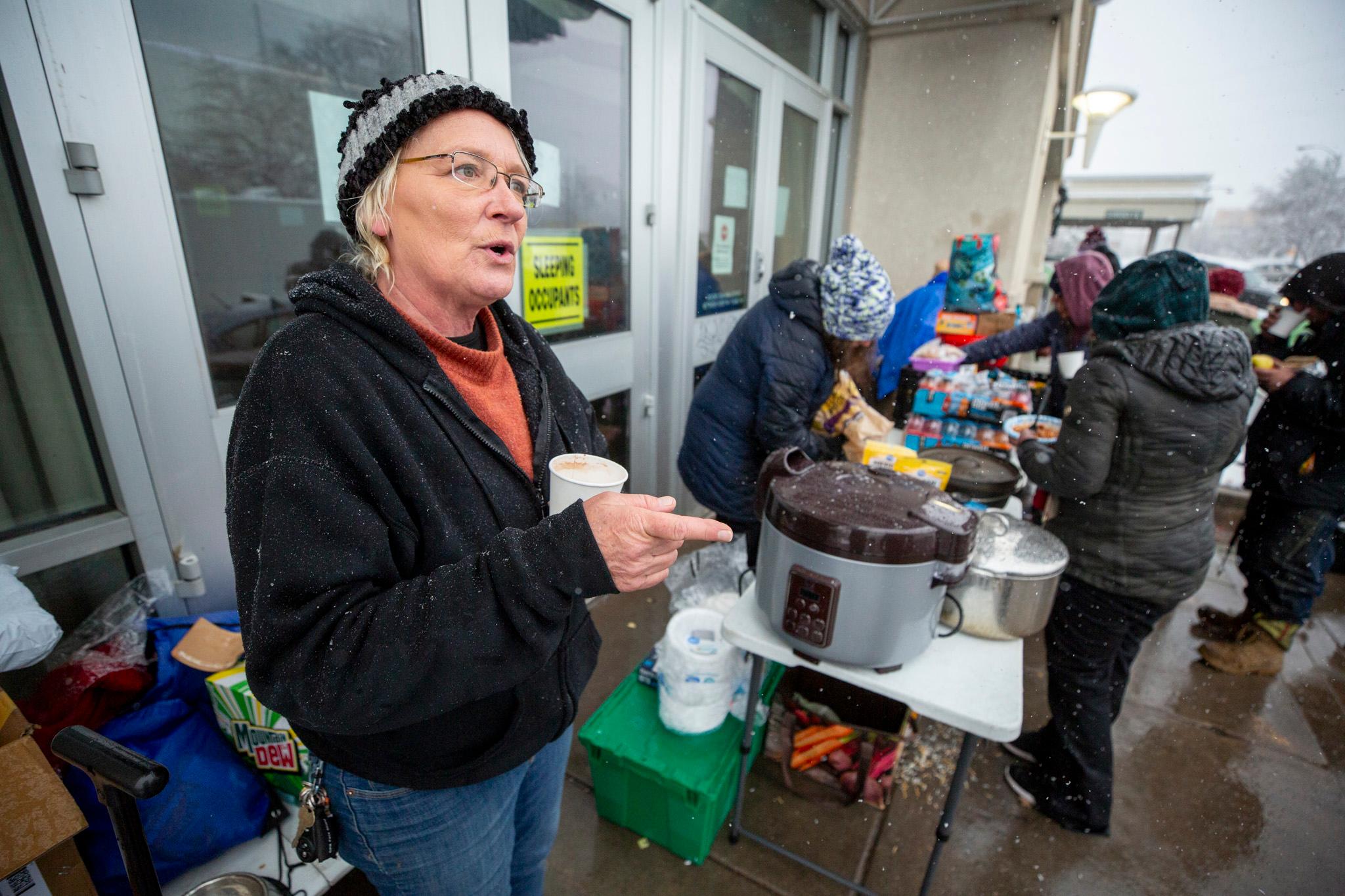 A woman in a winter hat holds a cup of coffee as people gather at a table covered with food and supplies behind her.