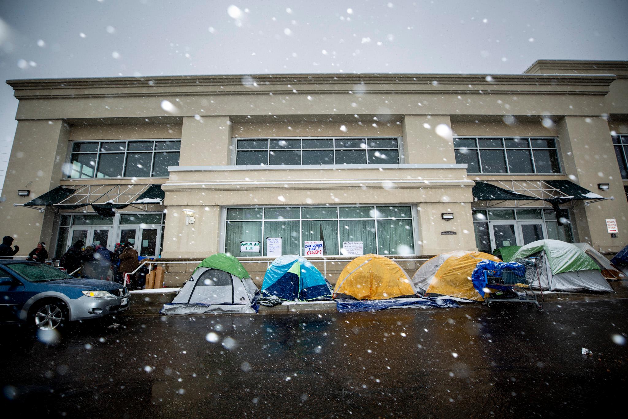Colorful tents are set up in front of a beige building as snow falls in the foreground.