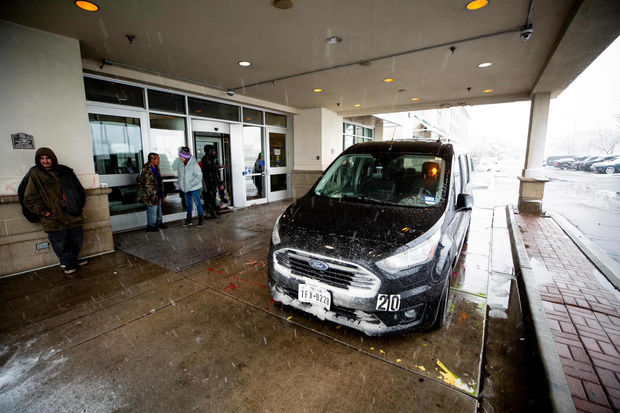 A black van is parked under a hotel overhang. Snow falls in the foreground as people mill around by the door.