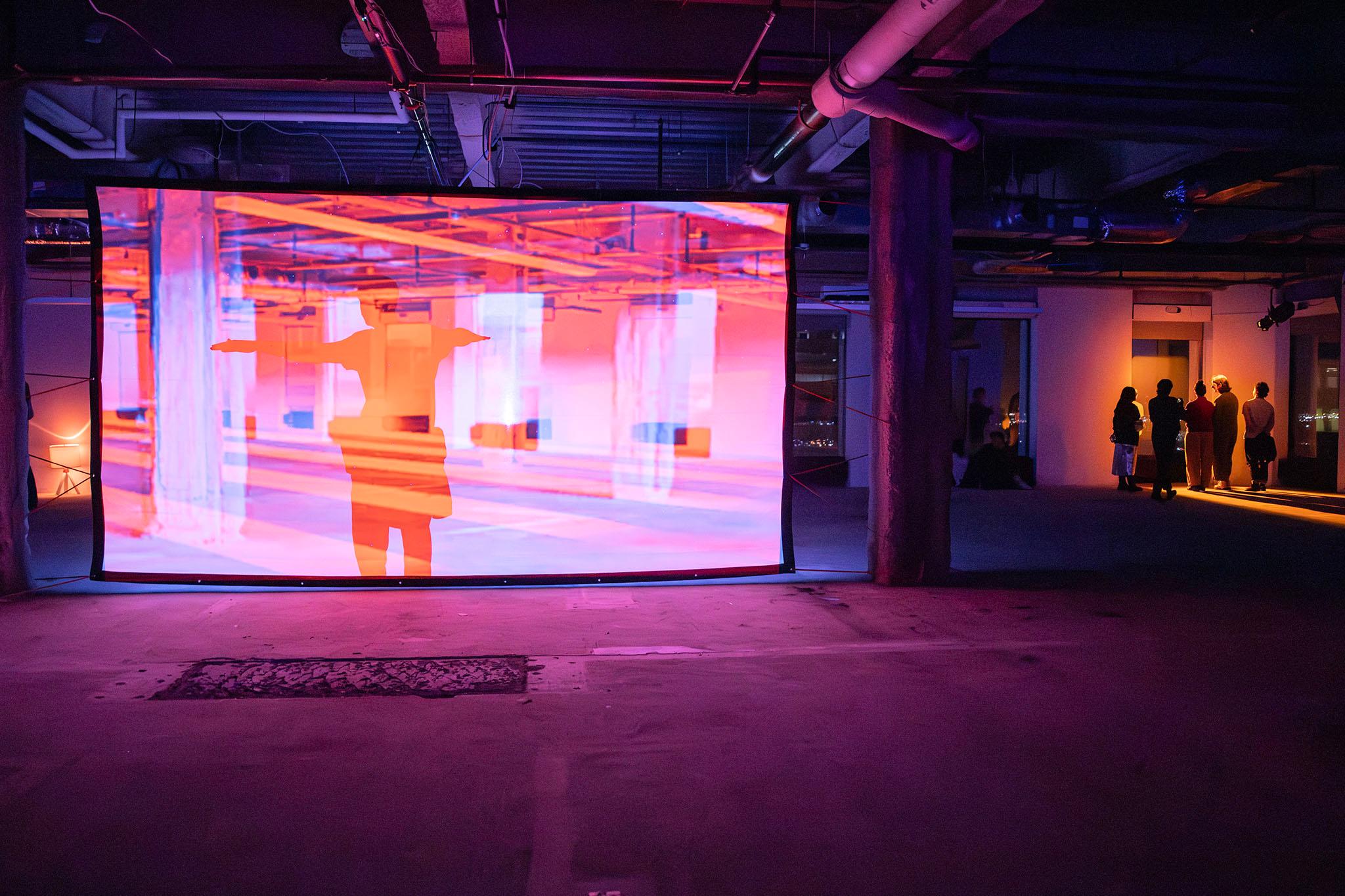 A projected screen sits in a bare space with concrete floors. People stand in a wash of orange light behind it.