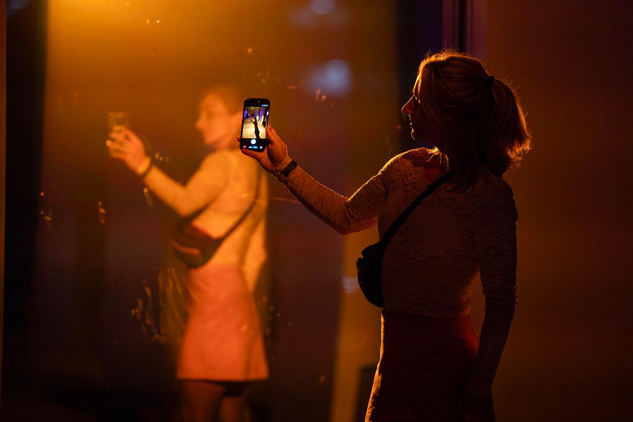 A woman dramatically lit by an orange light points a camera at a piece of reflective glass, showing the other side of her.