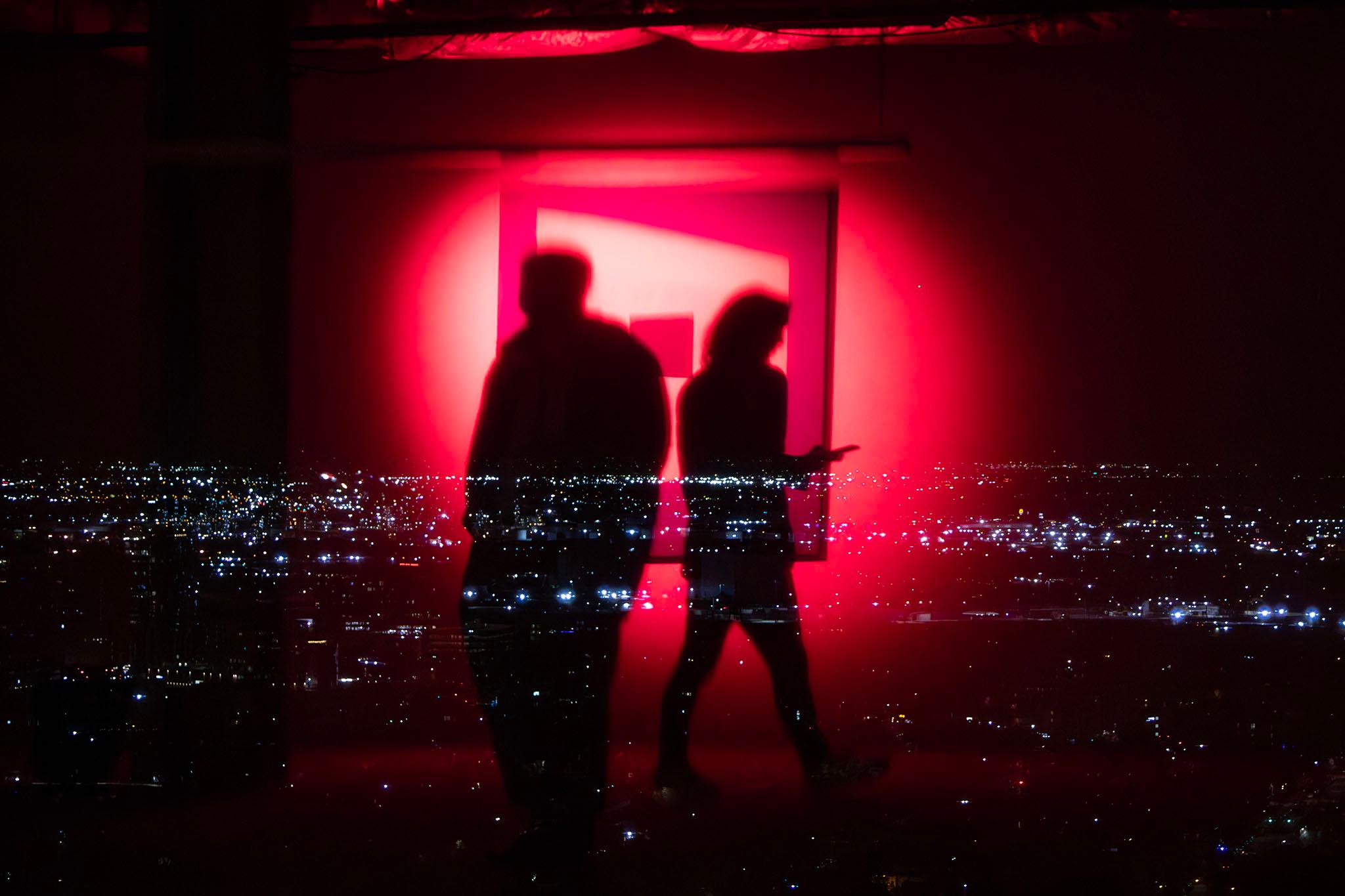 A view out of a tall building shows the lights of a city stretching into the distance. On top of it is the reflection of two people silhouetted by a glowing red light.