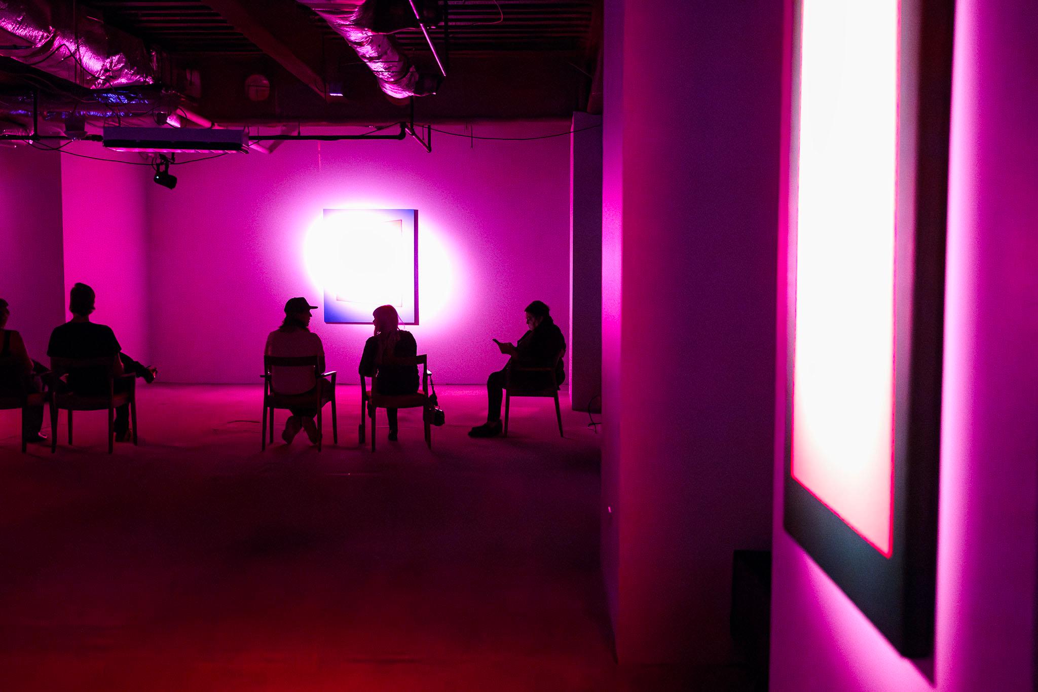 People sit in chairs, silhouetted by pink light washing over the walls. The space is otherwise bare, with concrete floors and shiny HVAC equipment mounted in the ceiling.