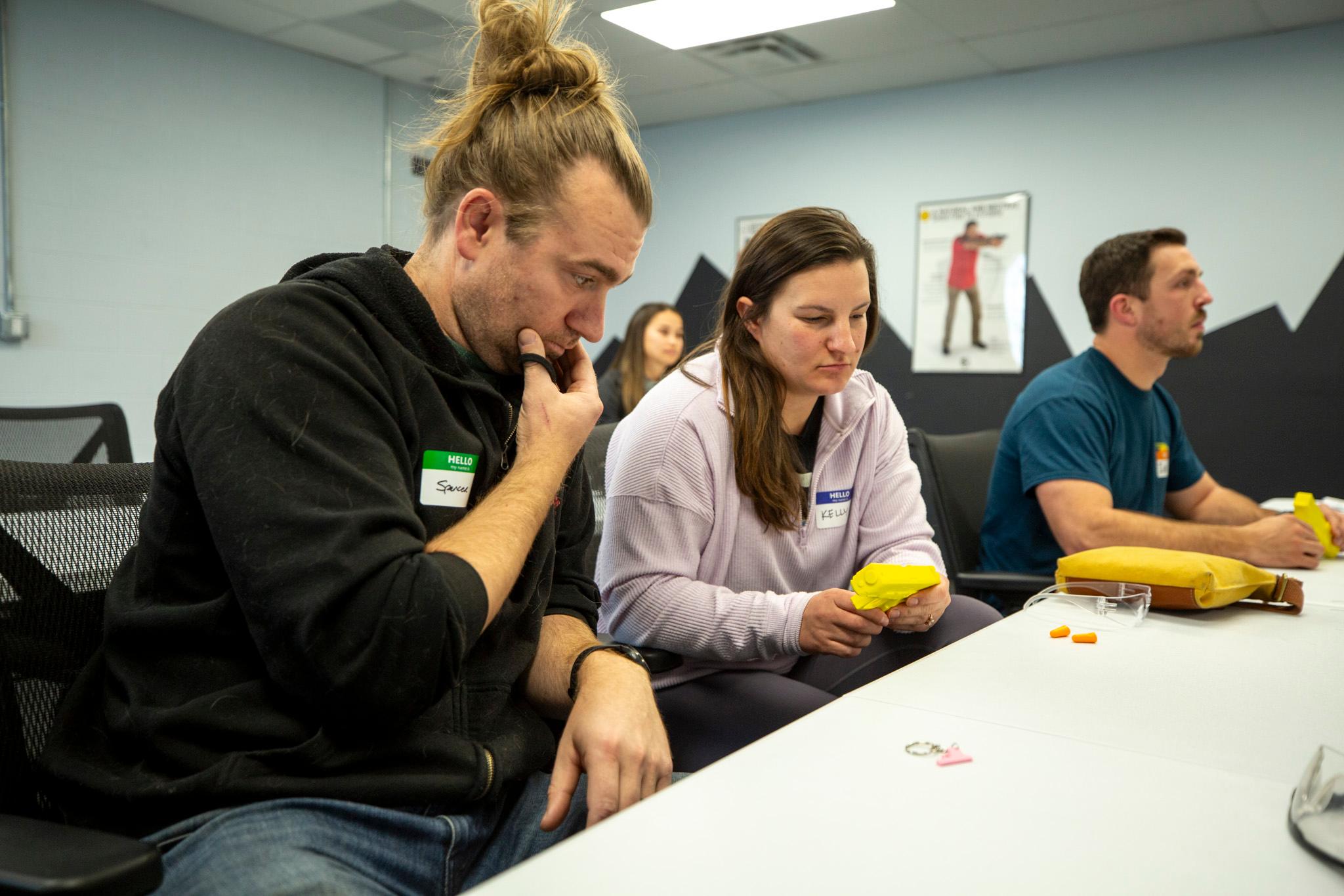 A man and a woman look down at a yellow, plastic pistol at a classroom table.