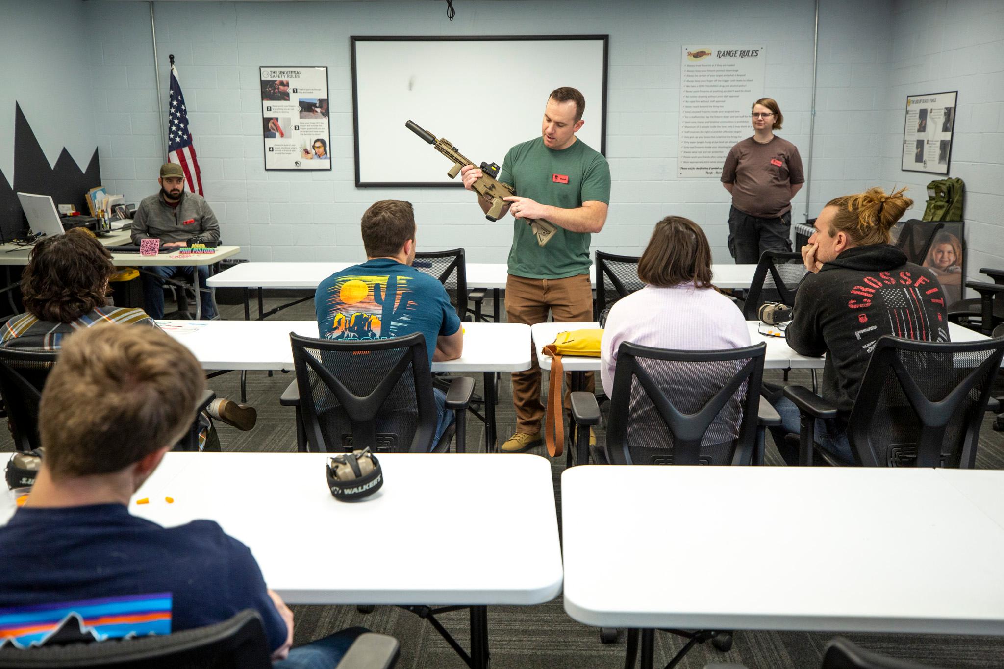 A man holds a green rifle at the front of a classroom as people watch.