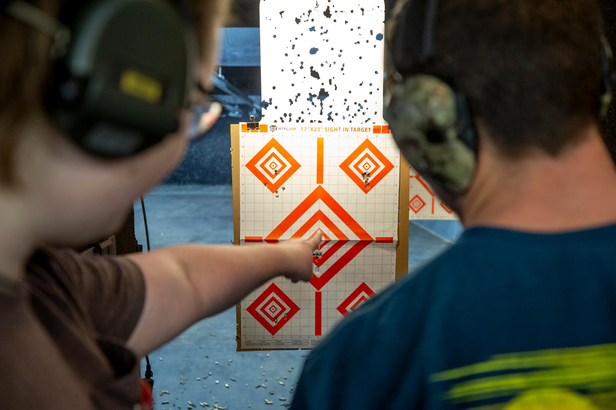 Two people stand in the foreground, each wearing ear protection, as the person on the right points to a target full of holes.