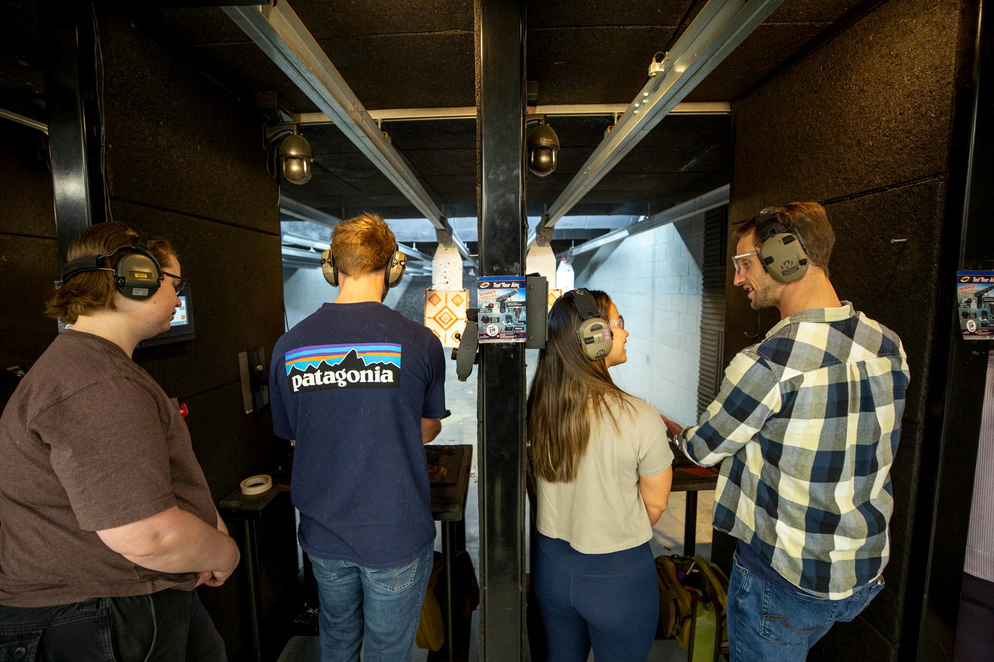 Four people wearing beefy ear protection stand in two black booths with targets hanging in the distance.