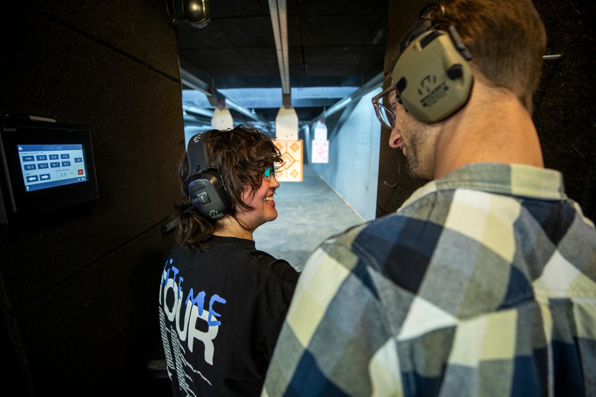 A woman and a man, both wearing beefy ear protection, look at each other in a black booth with targets hanging in the distance.