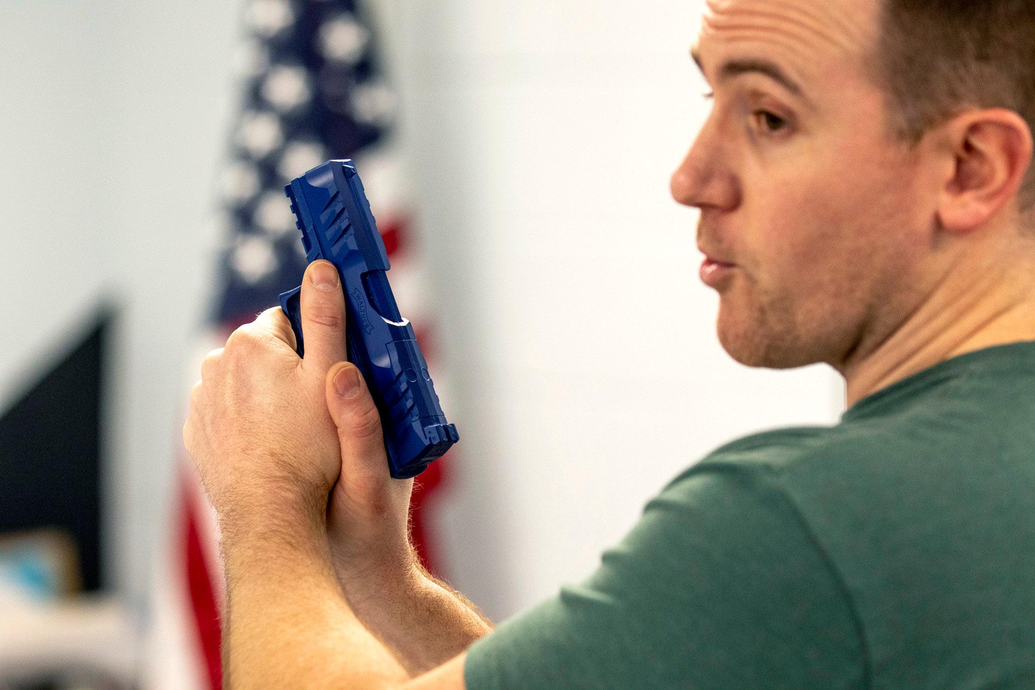 A man in a green shirt holds a blue plastic pistol. An American flag sits in the distance.