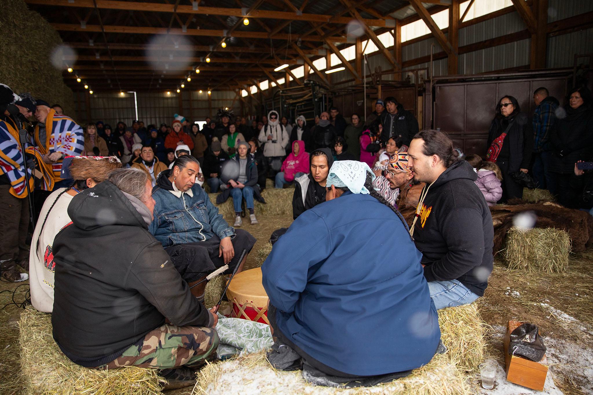 A group of men sit on haybales around a red and tan drum. The large barn they sit in is filled with people watching.