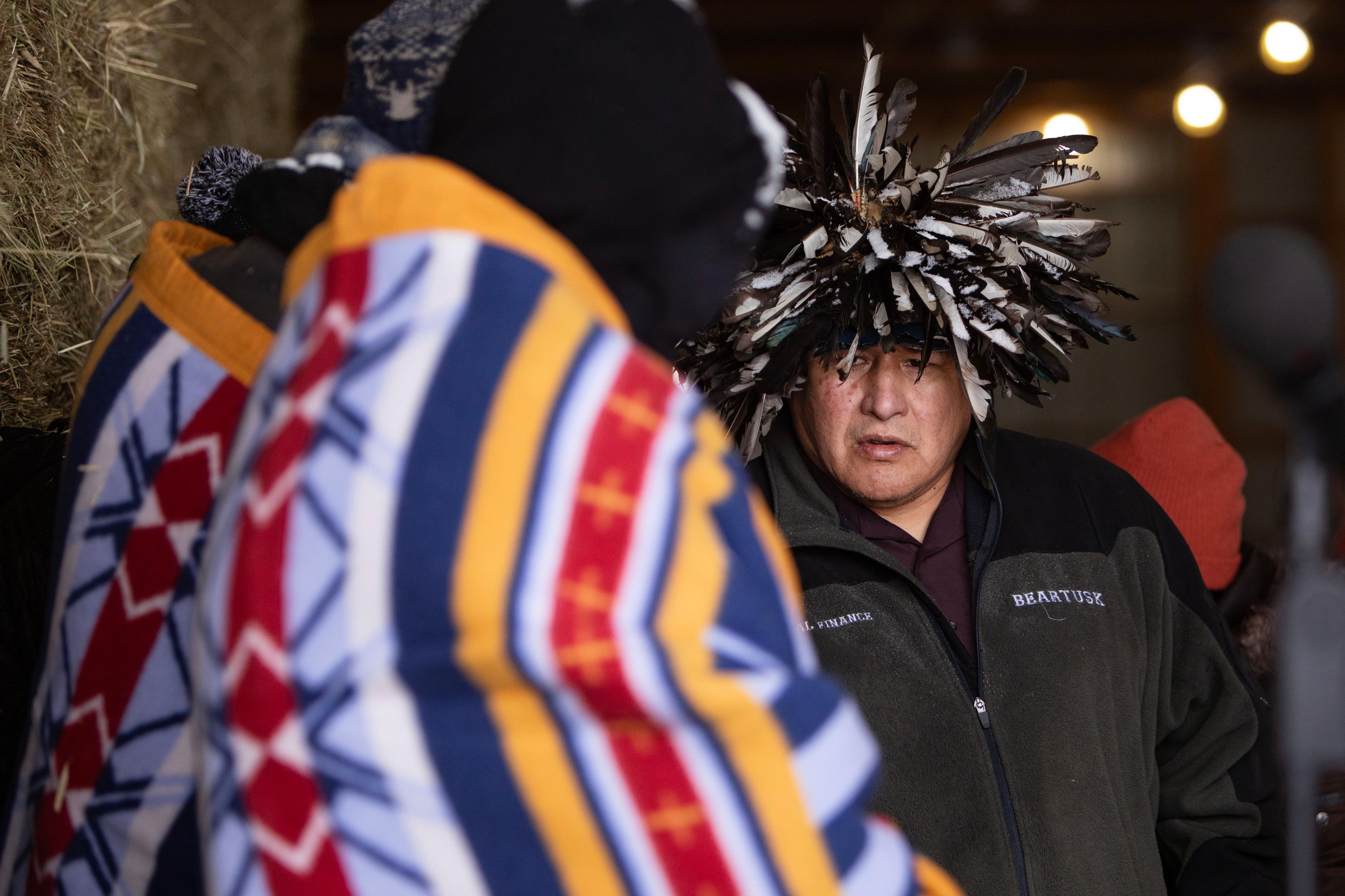 A man wearing a hat of black and white feathers stands before another person wrapped in a red, blue, yellow and white blanket.