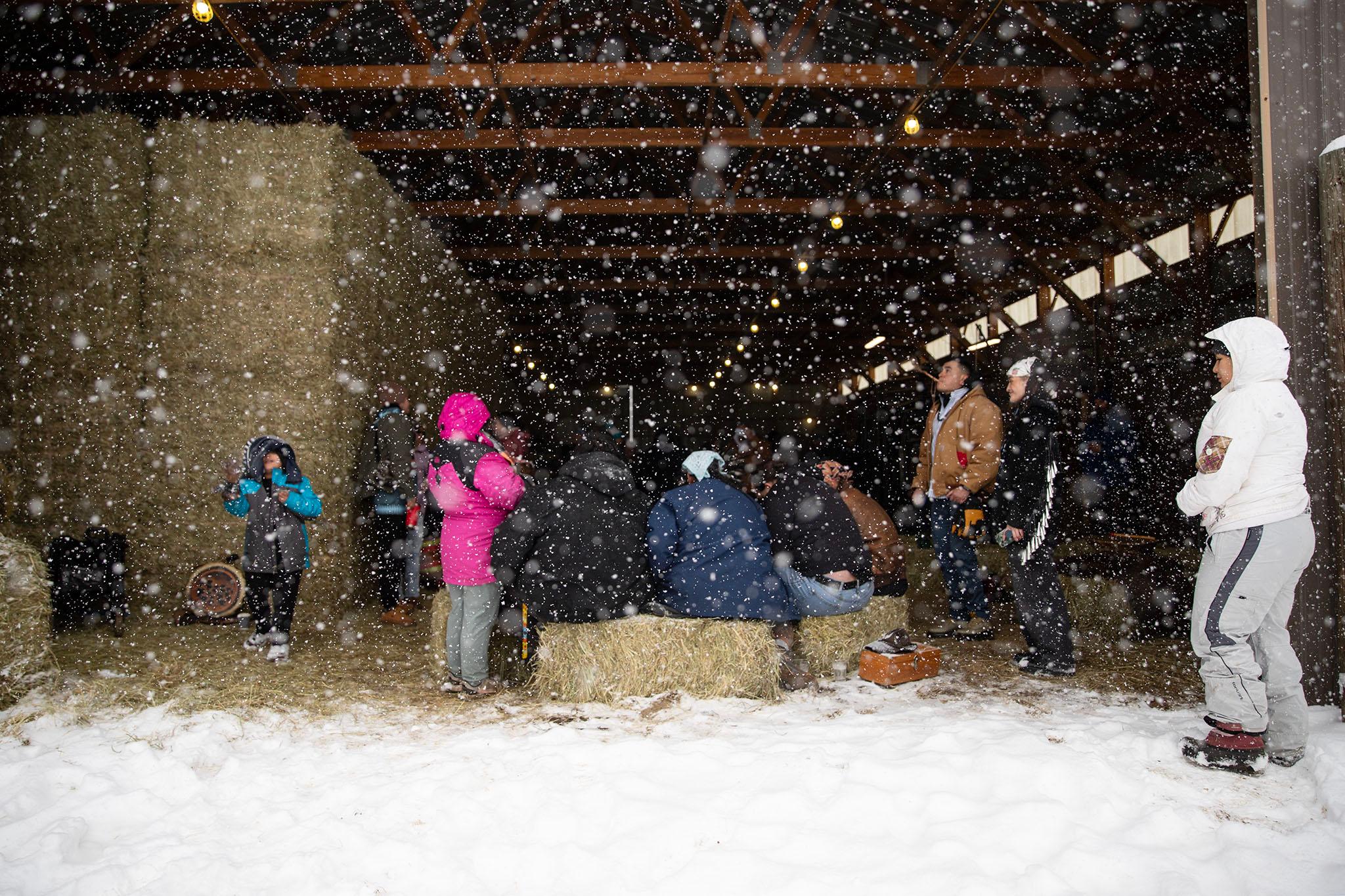 People stand inside a barn, sitting on haybales and milling around, as snow dumps in front of the door before them.