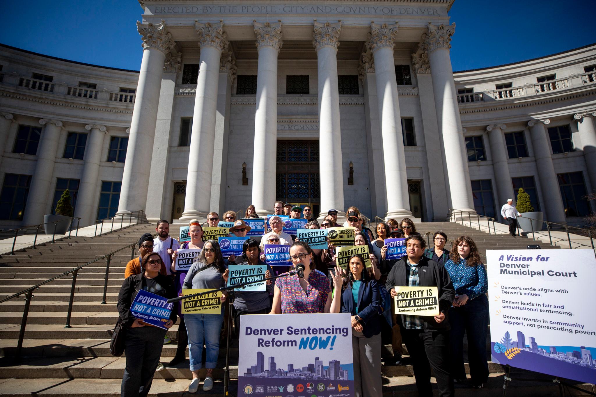 A woman in a colorful shirt speaks into a podium microphone as a crowd stands on stone steps behind her. They hold signs that read, "Poverty is not a crime!" A stone, columned building fills the background.