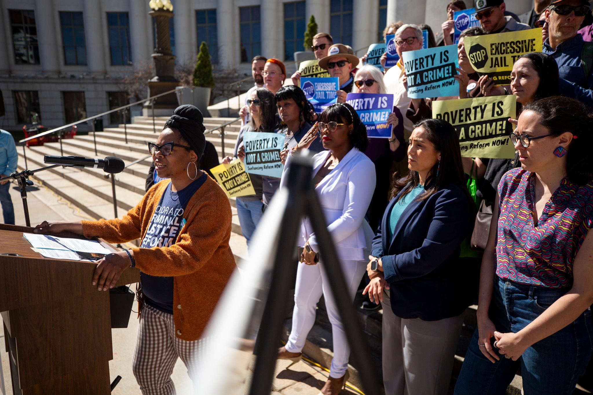 A woman in an umber sweater speaks at a podium microphone as a crowd stands on stone steps behind her. They hold signs that say, "Poverty is not a crime!"