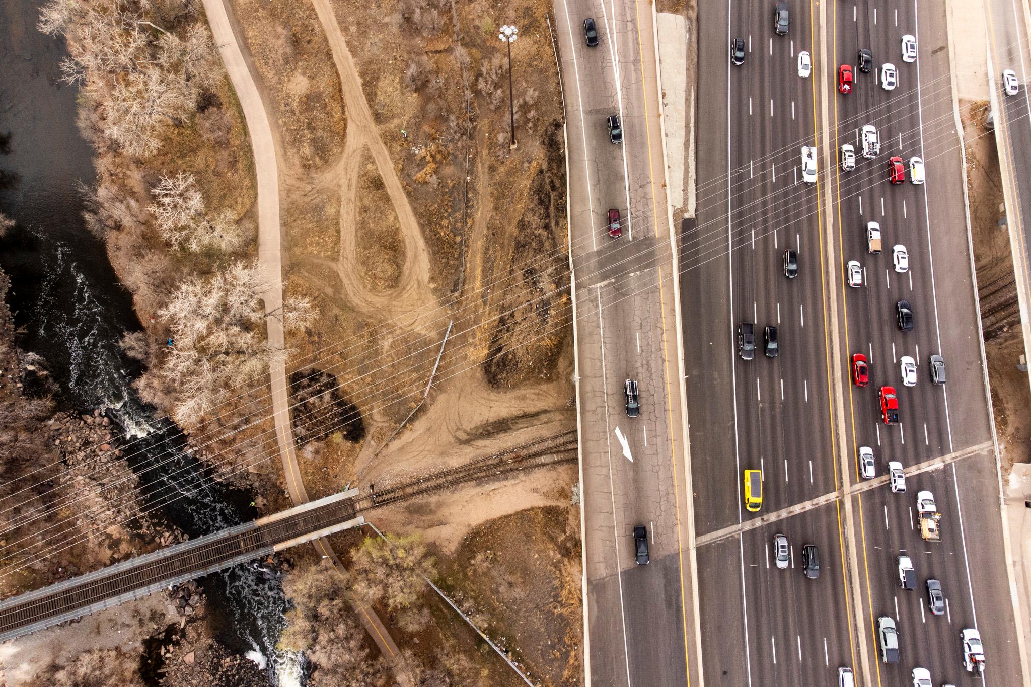 An aerial view of brown dirt, on the left, and a many-laned highway, on the right. Power lines and a railroad track stretch across the frame. A river flows on the left edge.