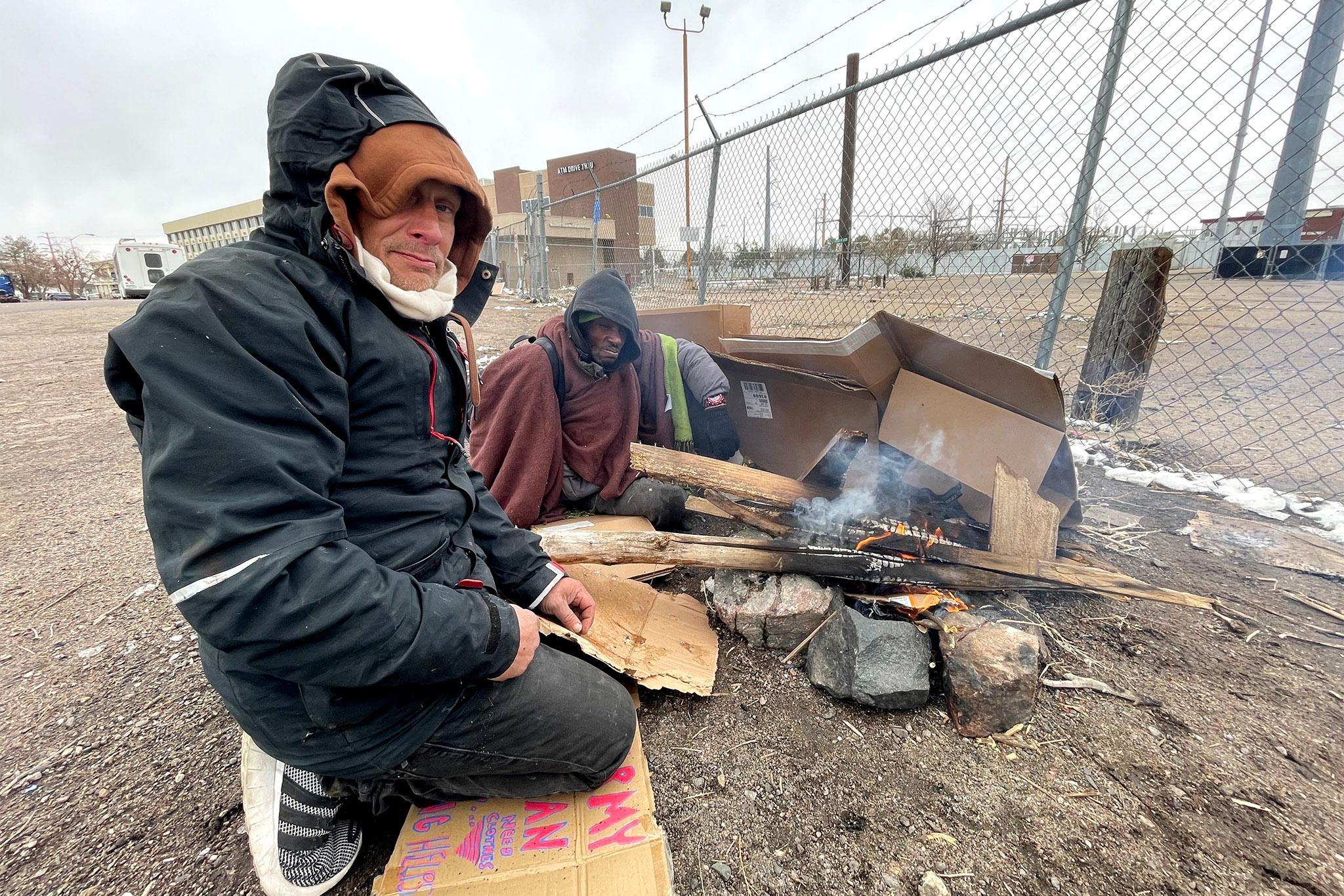 Two men, bundled up in hooded jackets and blankets, sit on a dirt ground around a fire made with some pieces of broken wood, cardboard and rocks. A barbed-wire fence stretches through the frame.