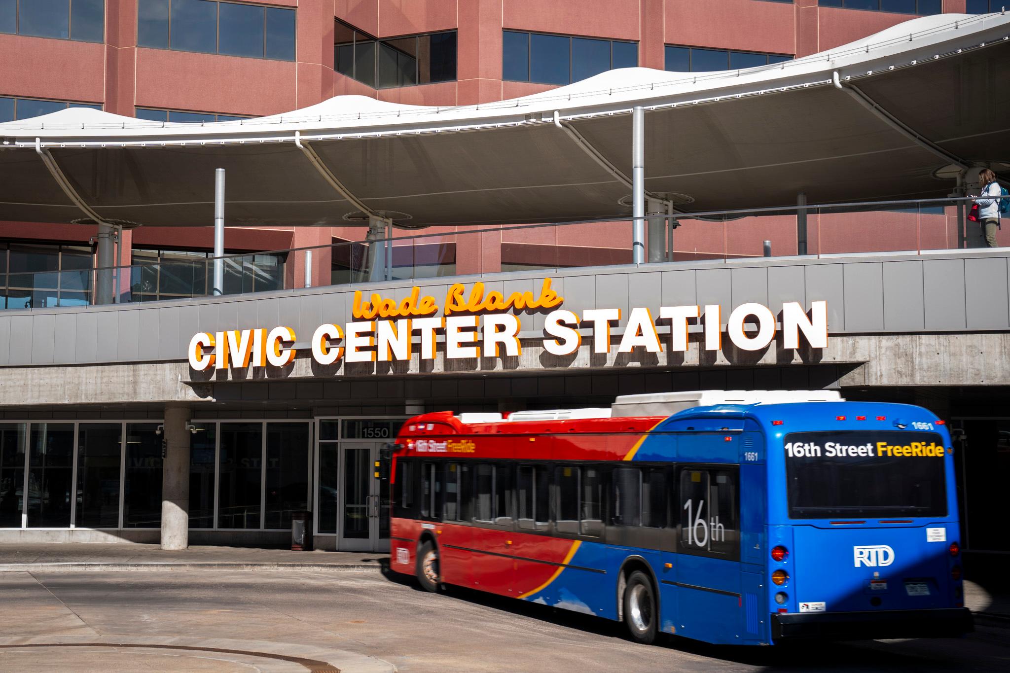 A red and blue bus stands before a concrete pavilion, where lettering hung above a row of doors reads "Wade Blank Civic Center Station" in yellow cursive and large white sans-serif fonts.