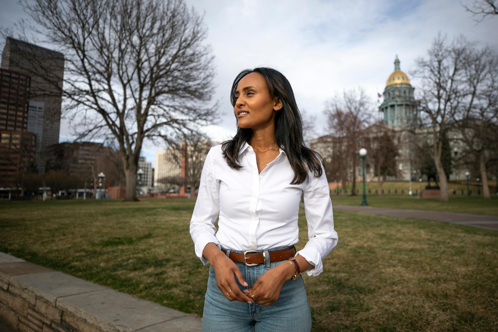 A woman in a white button-up stands in front of a park of green grass and leafless trees. A gold-domed building rises in the background.