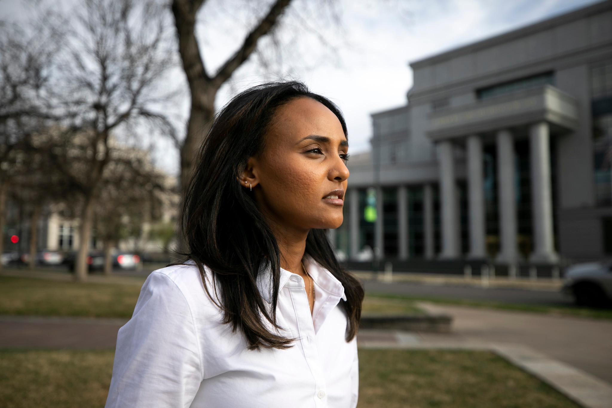 A woman in a white button-up stands in front of a park of green grass and leafless trees. A building of stone columns rises in the background.