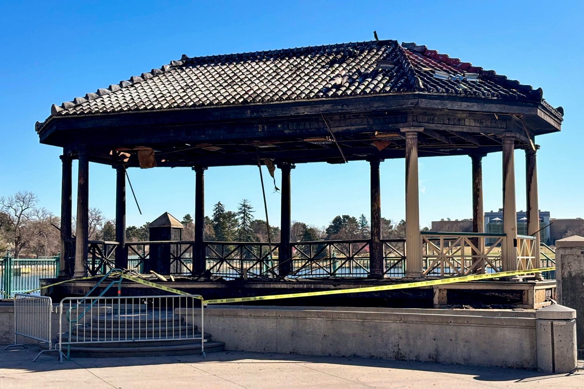 A roof of tiled shingles, atop a set of columns, is black with fire damage. A strand of yellow police tape is wrapped around it, and a metal barricade blocks the steps into it.