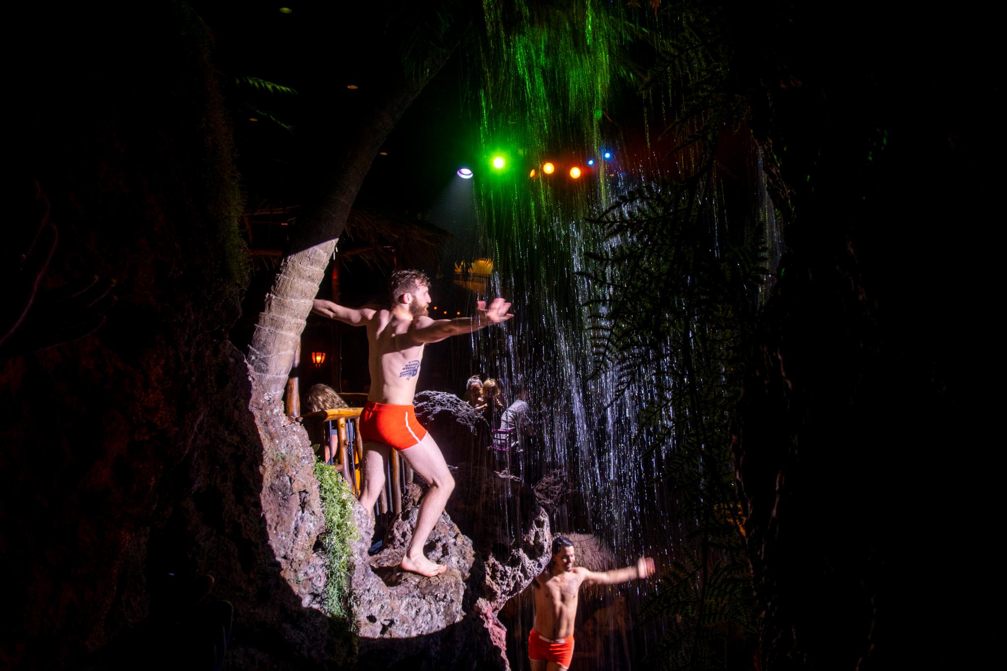 Two men in short red swim trunks stretch out their arms as they perch on rocks beneath a waterfall.