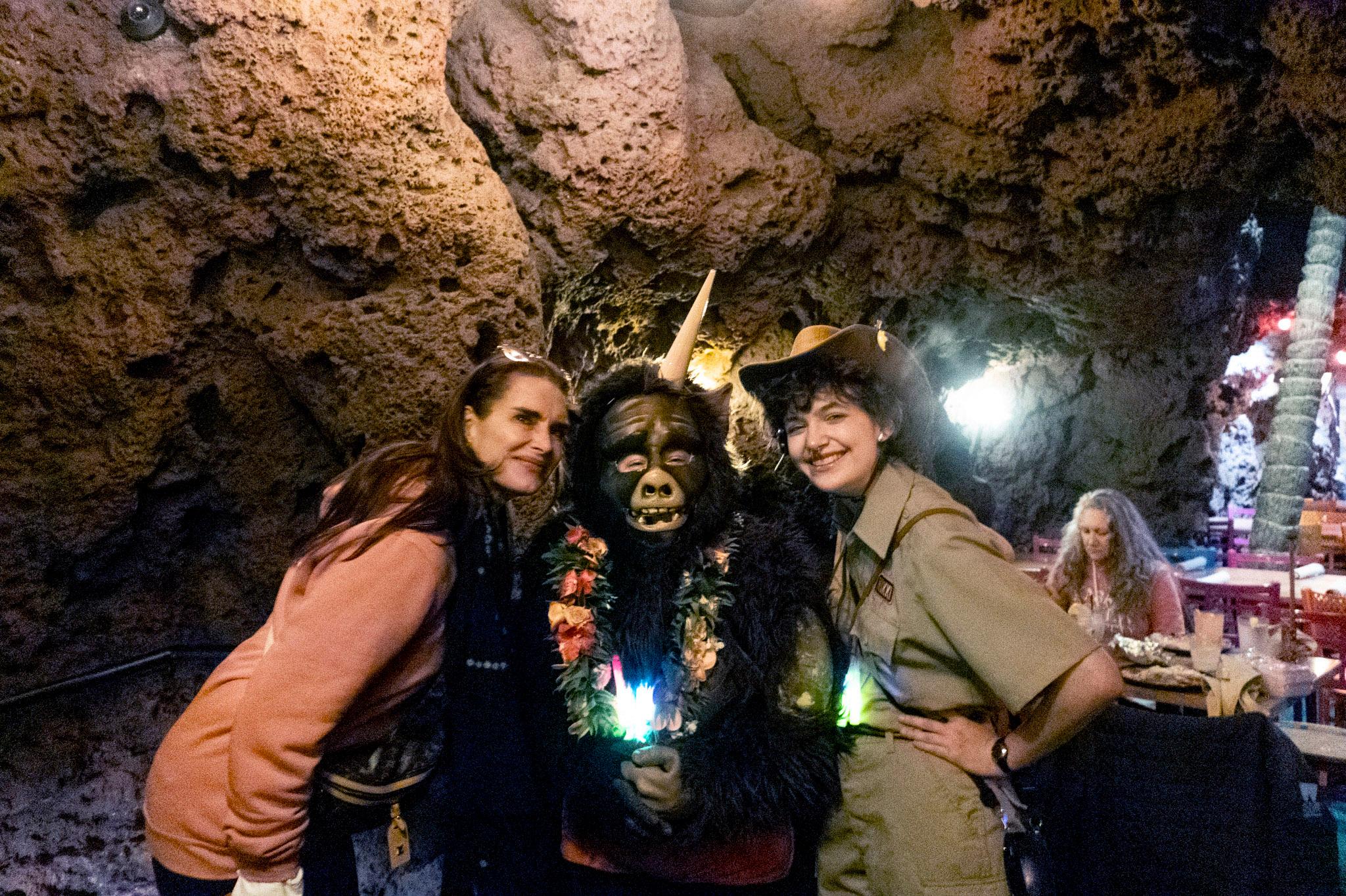 A woman in a pink sweatshirt poses with someone in a gorilla costume and someone else dressed like a safari guide, in a room outfitted to look like a cave.