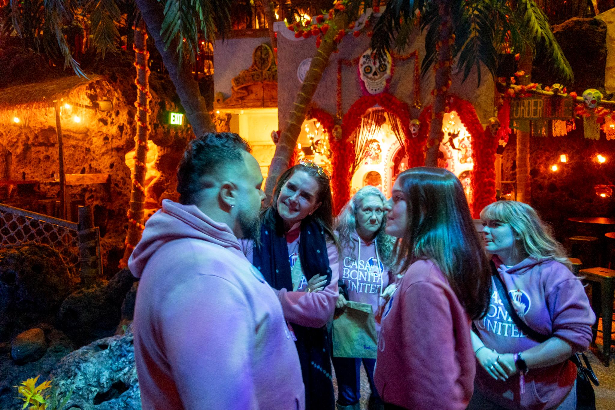 A woman in a blue scarf cocks her head as she speaks with a group of people in pink sweatshirts. They stand in a dark room with glowing lights, filled with palm trees and rustic-looking buildings. A smiling skull hangs over a doorway in the background.