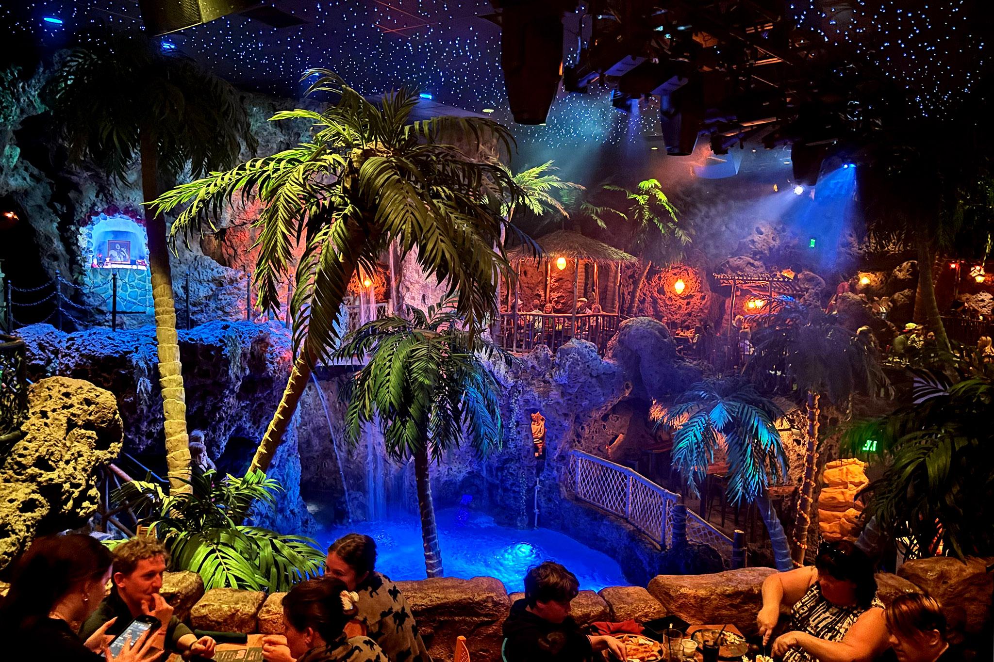 A glowing blue lagoon sits beneath palm trees and thatched-roof huts as people dine at tables. The ceiling is speckled with lights that look like stars.