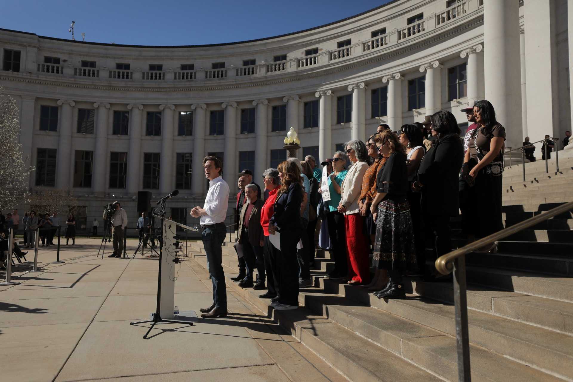 Photographed from the side, a crowd of people stand on the steps outside a white building with rows of columns and windows. A streak of blue sky is visible at the top of the photo.