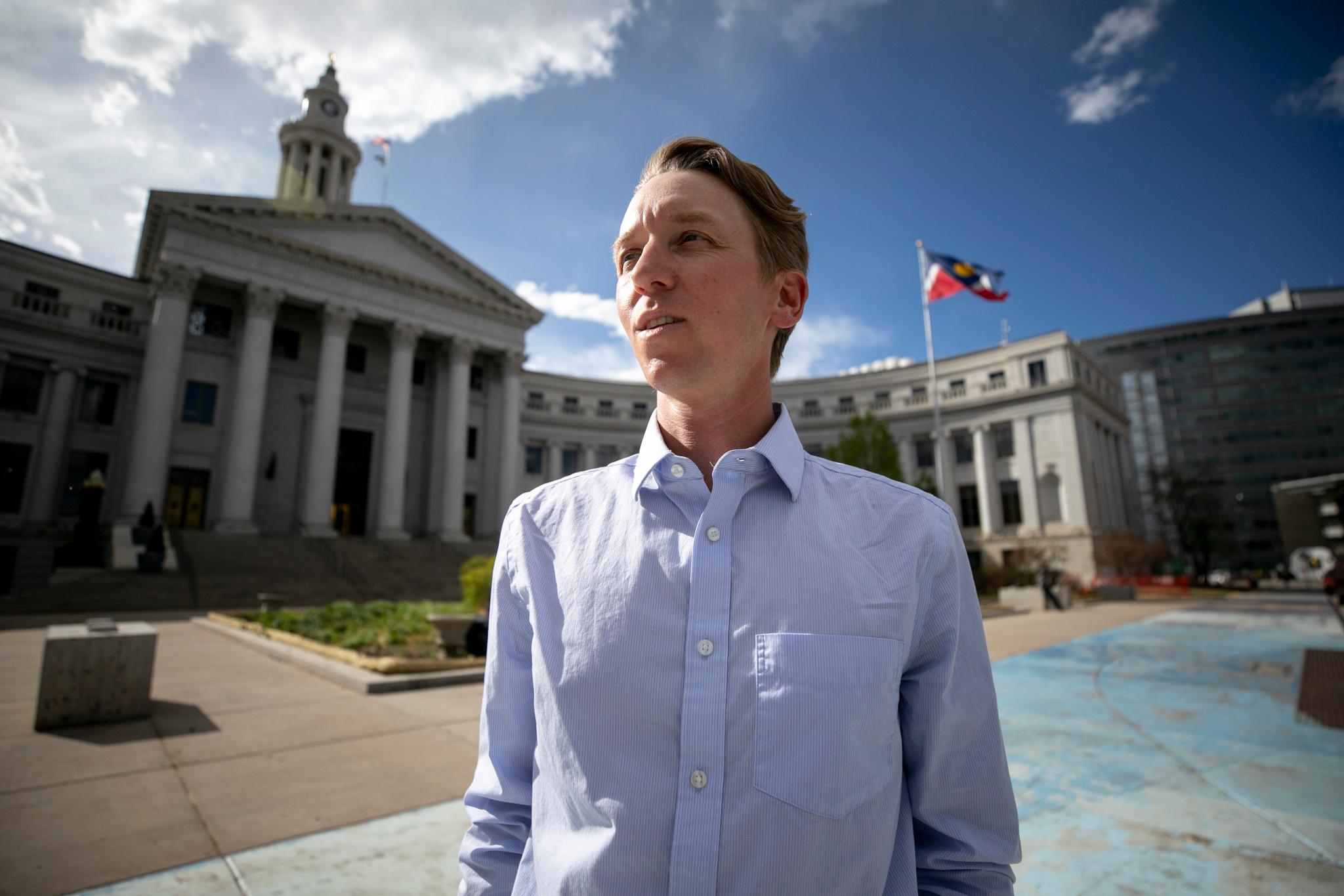 A man in suitpants and a button-up stands in front of a large stone building of tall columns. A flag waves above him.