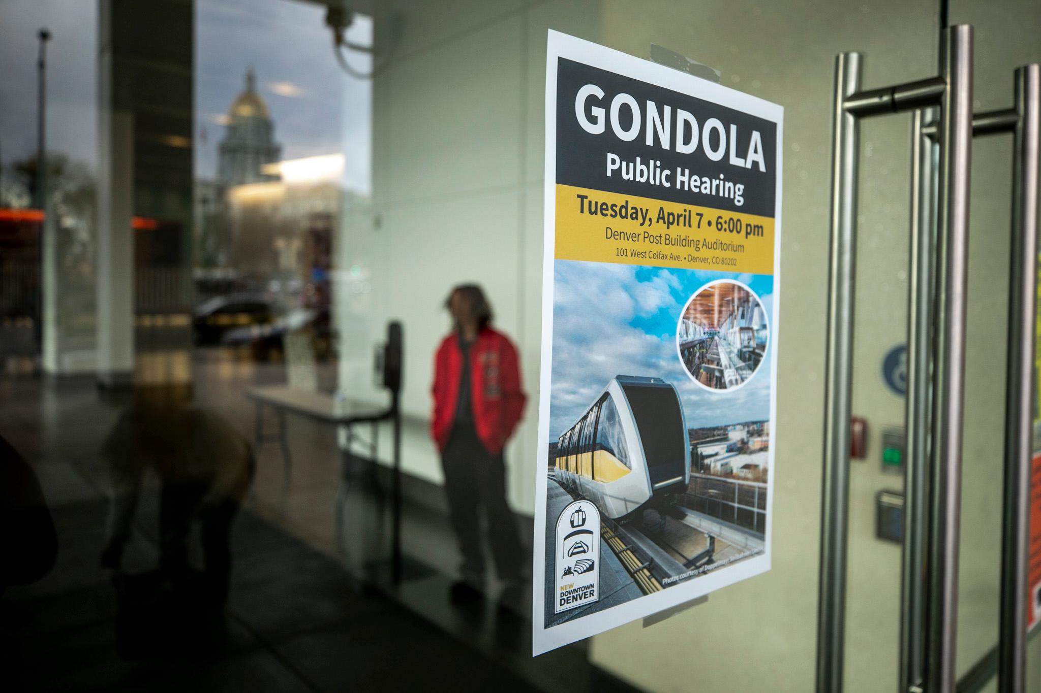 A poster on a glass door reads "GONDOLA Public Hearing" and features a photo of a train. The gold-domed Capitol building can be seen in the glass' reflection.