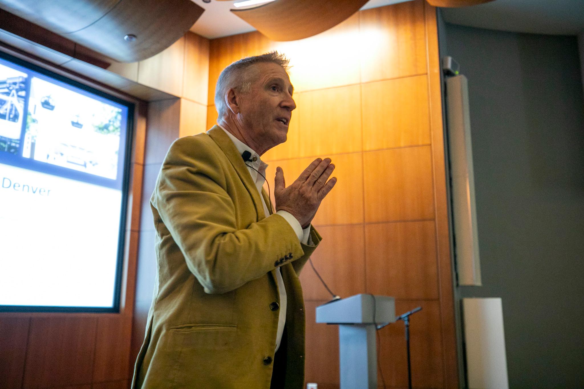 A man in a goldenrod jacket presses his hands together in a prayer-like pose as he speaks in a warmly lit auditorium.