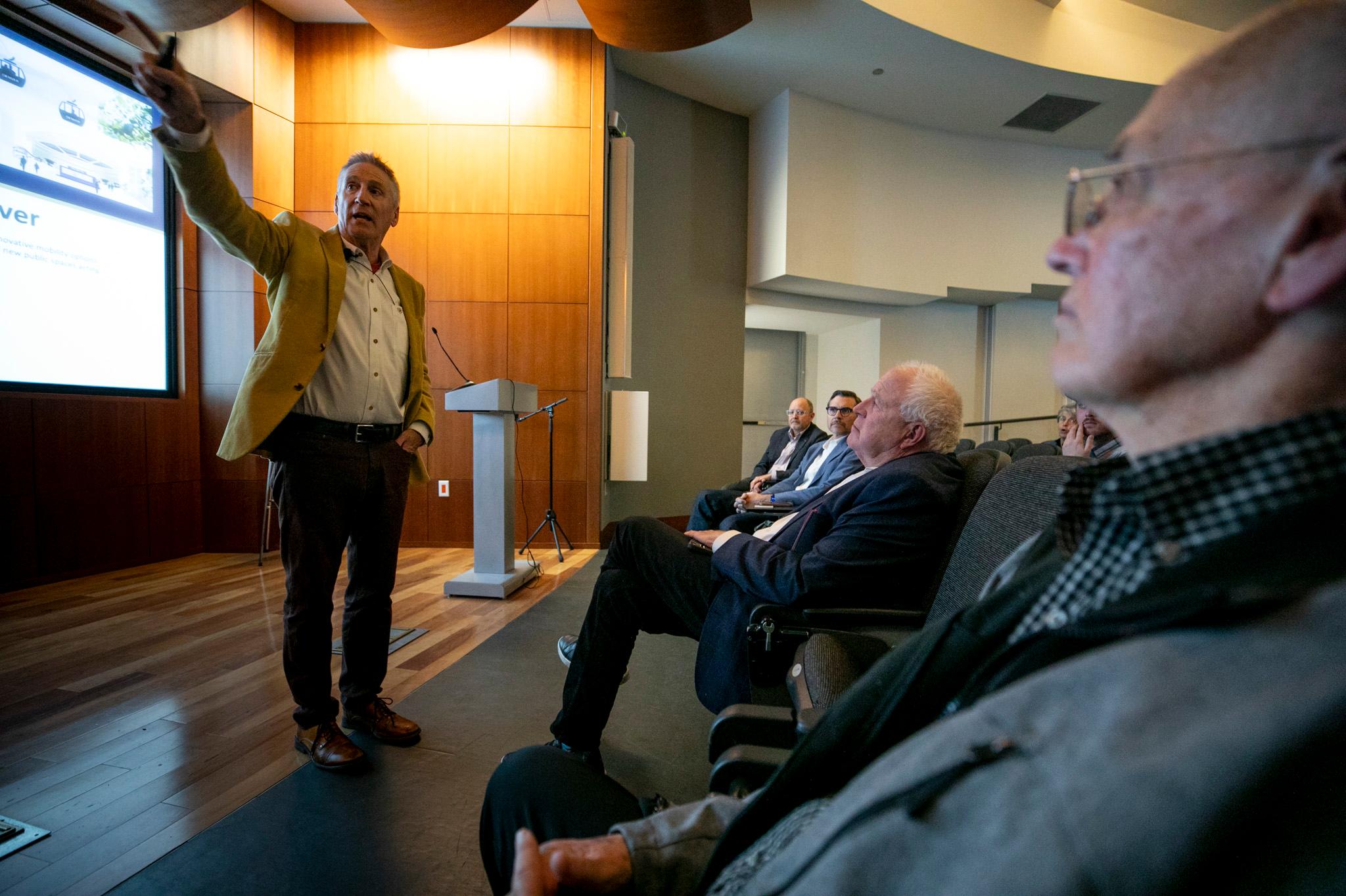 A man in a goldenrod jacket stands at the front of an auditorium, pointing up as he speaks.