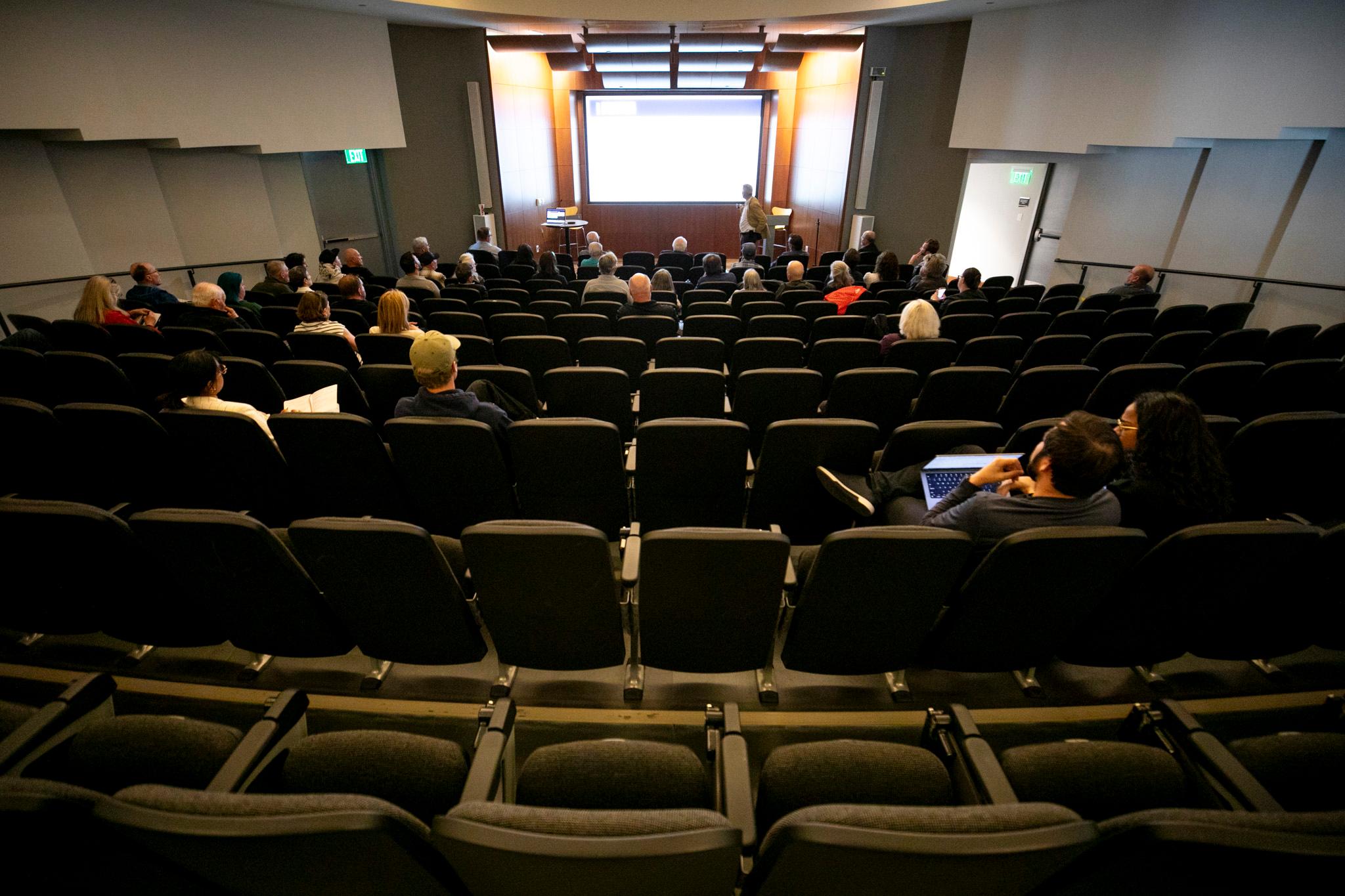 An auditorium is sparsely filled as a man in a goldenrod jacket speaks at the front, by a glowing white screen.