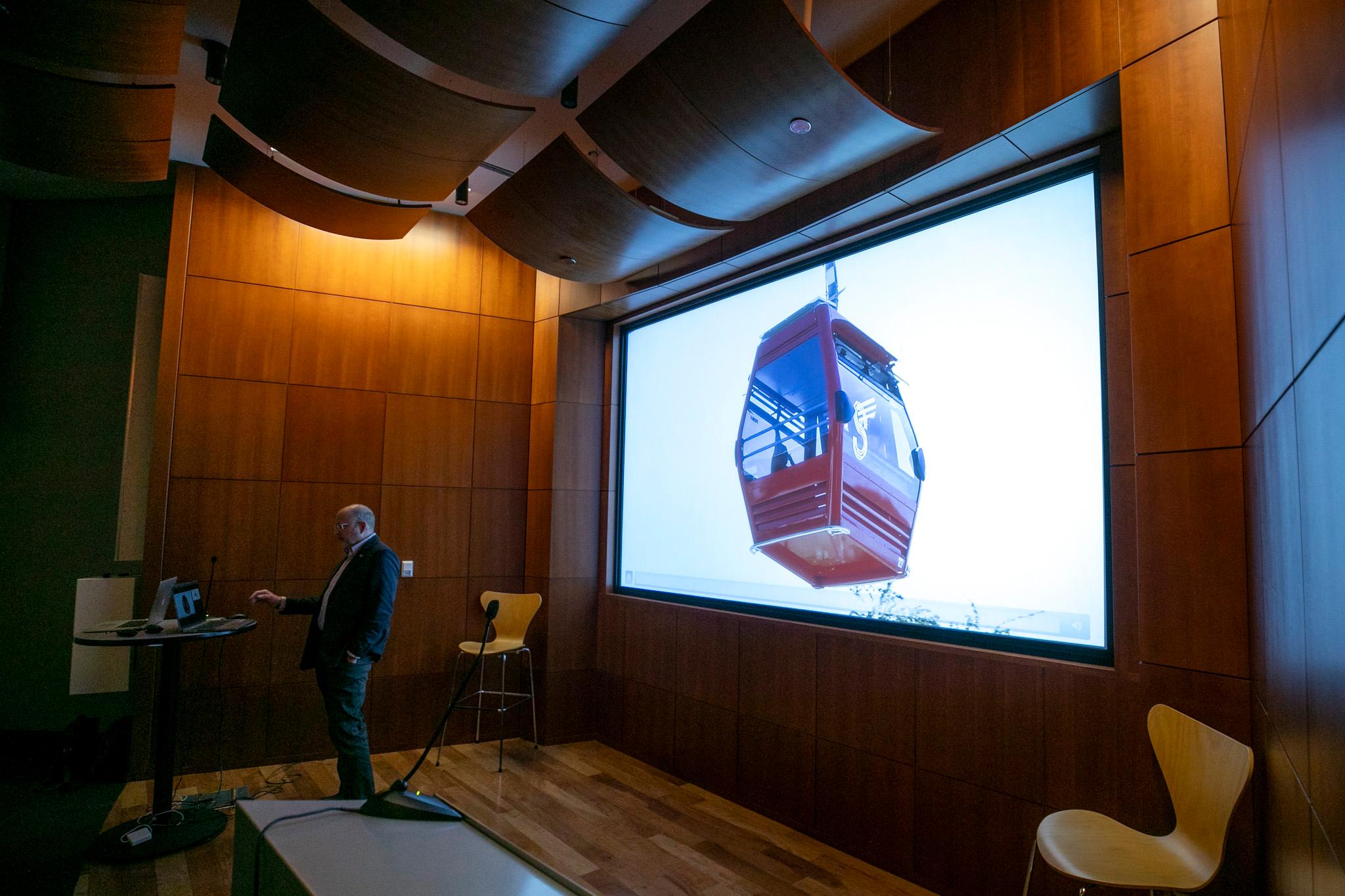 A photo of a red gondola car is projected on a screen as a man in a suit stands at a laptop before it.