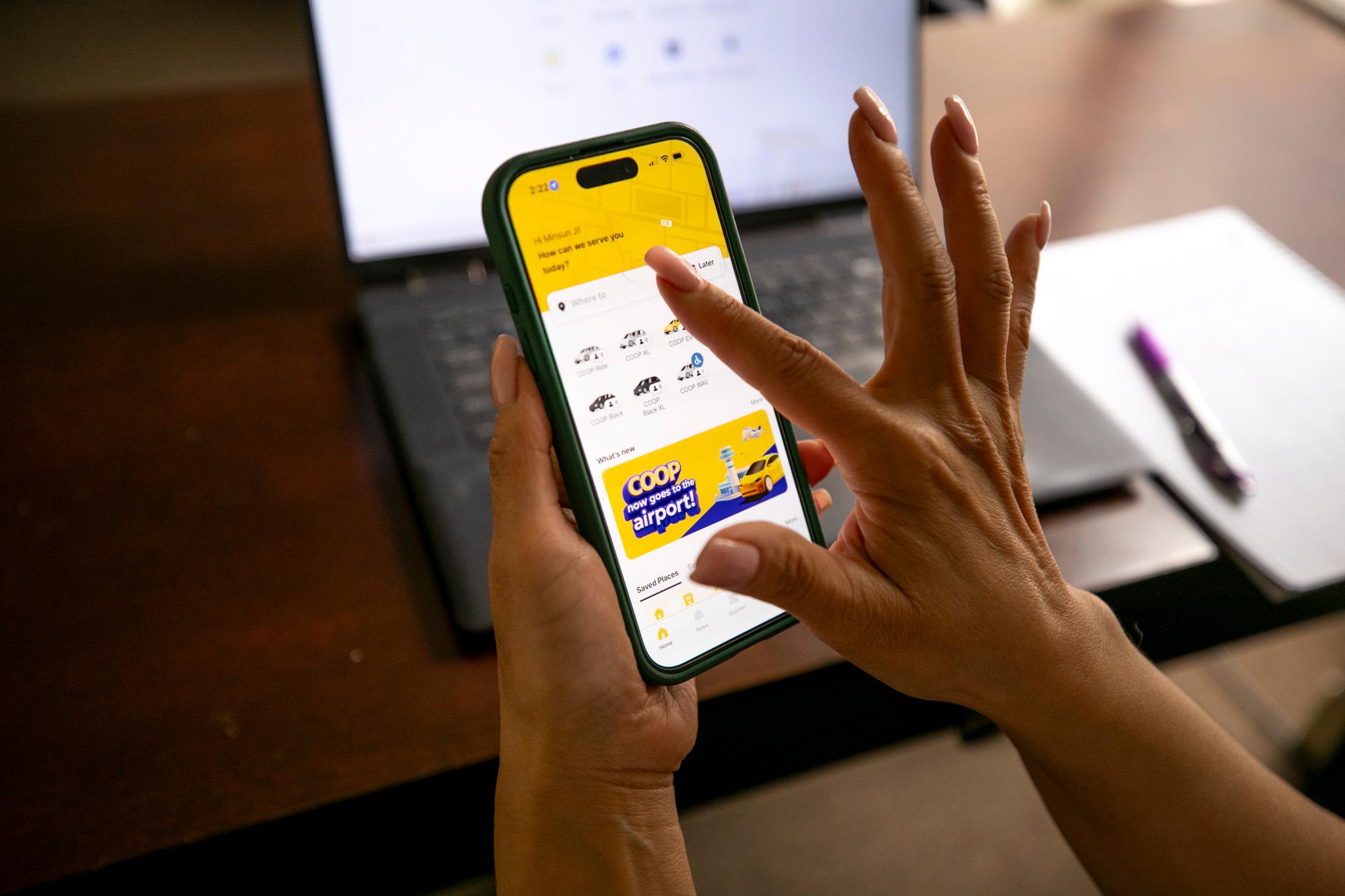 A woman's finger reaches to tap a smartphone, where a yellow and white app displays cars available for hire.