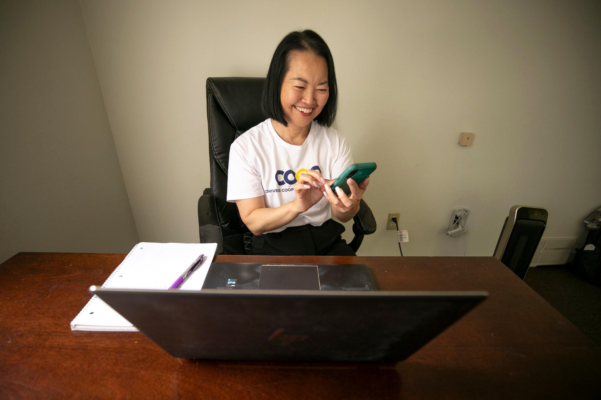 A woman smiles as she taps a phone, sitting at a wooden desk, behind a computer.