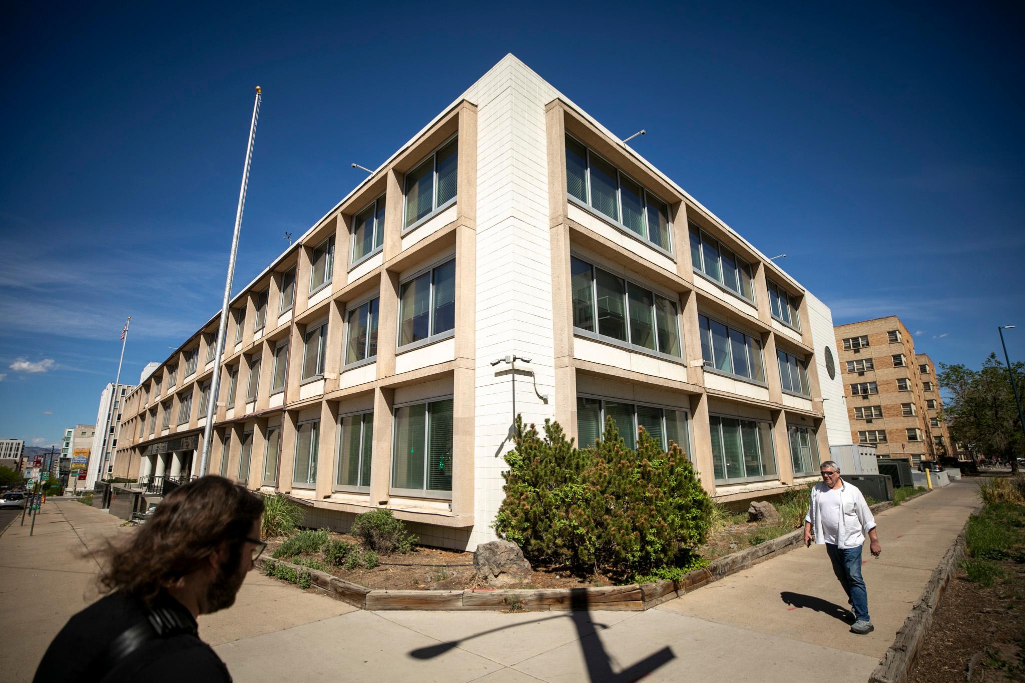 The corner of a boxy white and beige building. Two people walk by.