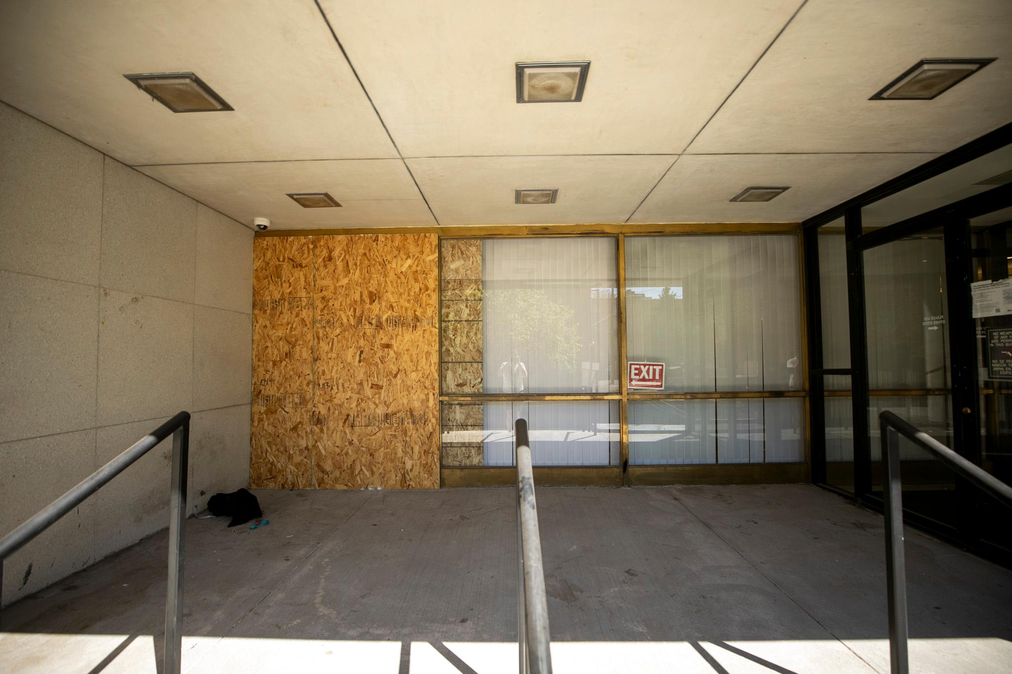 The glass-lined entrance to a building. One of the windows is covered in plywood.