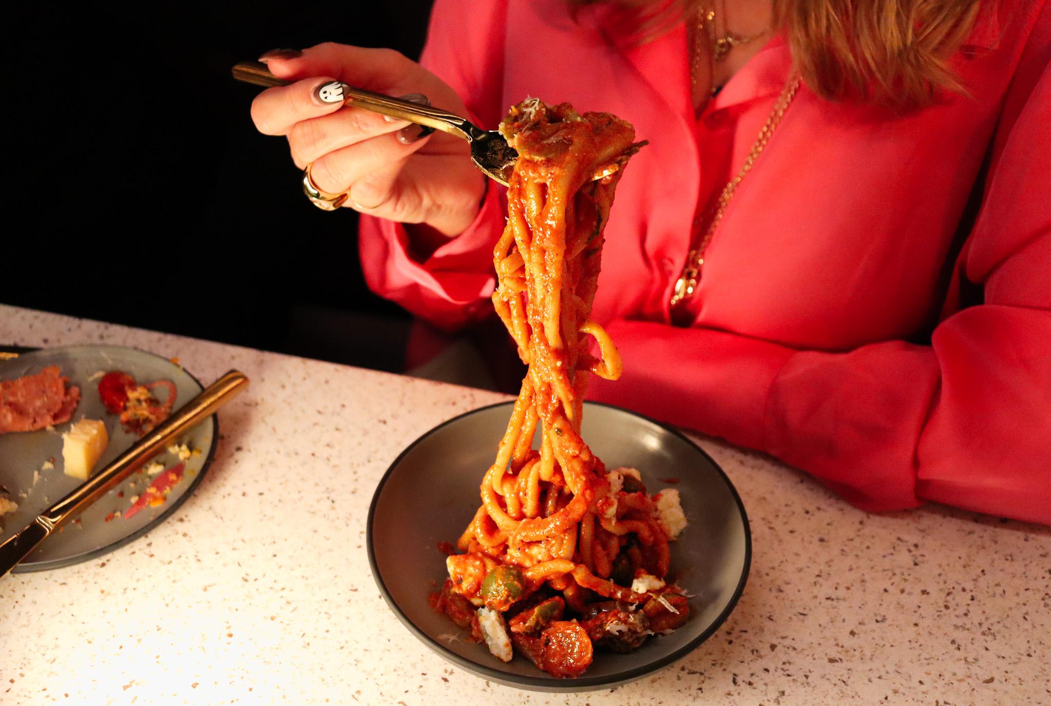 A person wearing a red button-up shirt lifts a big twirl of bucatini noodles covered in red sauce out of a grey bowl.