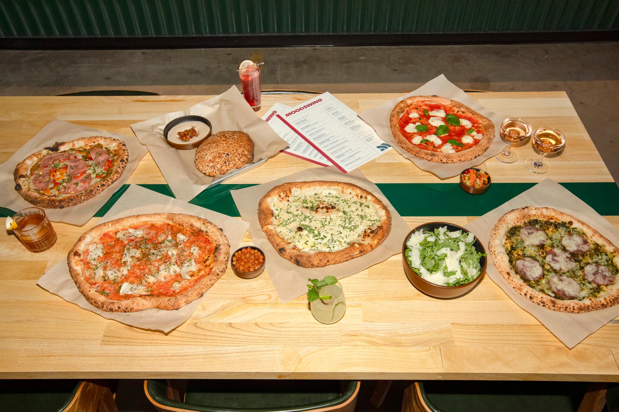 Five kinds of pizzas, a salad and a large piece of bread with a white bean dip are spread out on a wooden table. There are also a variety of drinks.