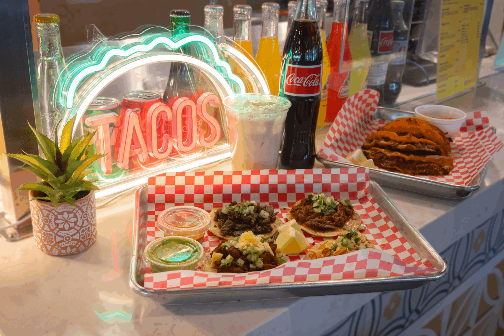 Two trays with food on red-and-white checkerboard paper are laid out on a white marble counter. The tray closer to the camera has four tacos, two kinds of salsa in plastic cups and a few lime slices. The tray further away has birria tacos. Behind the trays are a Mexican Coke bottle, a cup of horchata and a neon sign that says "Tacos."