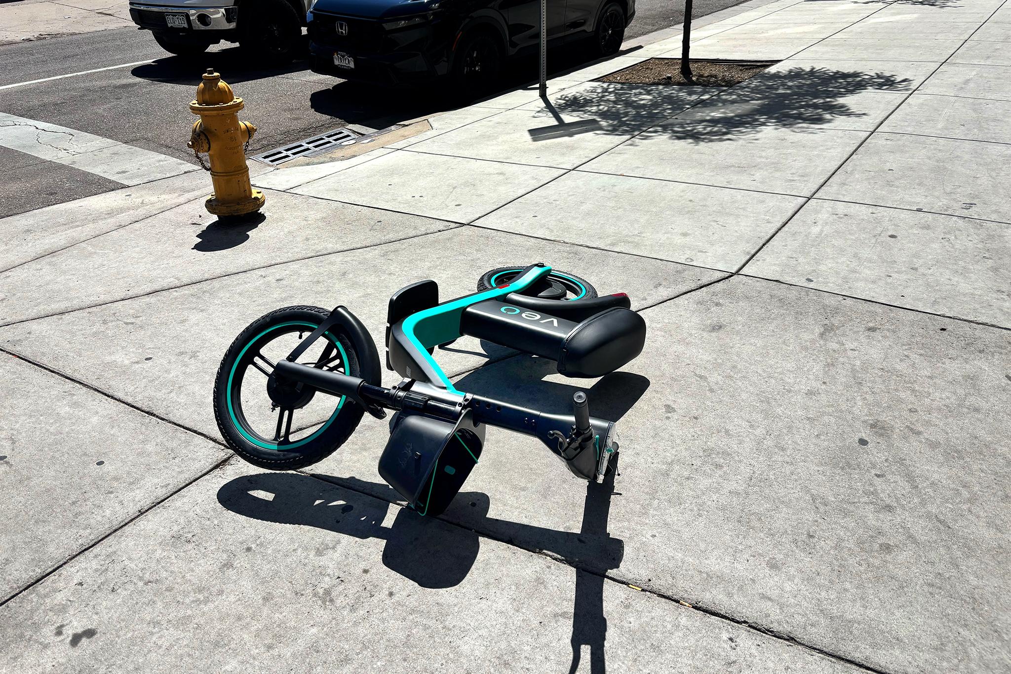 A black scooter with teal accents lies sadly on the sidewalk, as if in defeat.