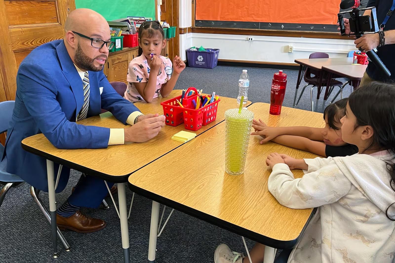 A man in a blue suit sits at a child-size table with four young girls, listening intently to them in their classroom.